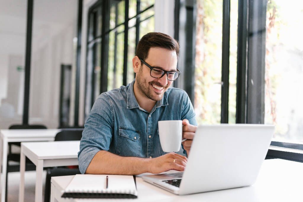 A laughing man looking at his laptop and holding a cup of coffee