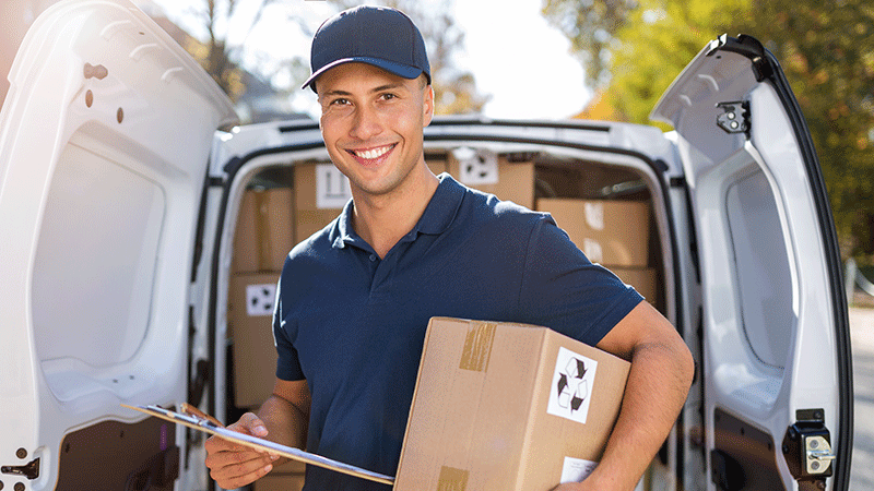 Smiling worker holding a box, standing behind his open van filled with more boxes.