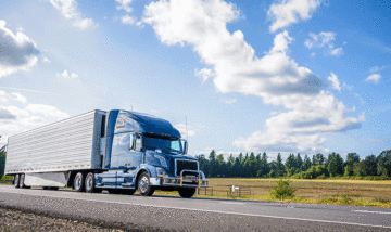 Truck driving down highway with clear blue skies and a field in the background