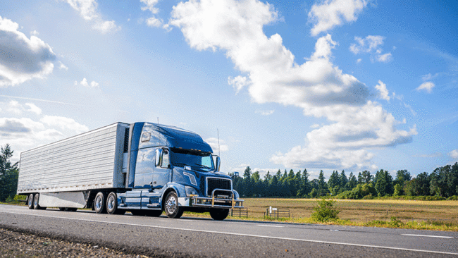 Truck driving down highway with clear blue skies and a field in the background