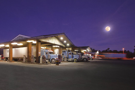 Outside of the Little America truck stop, trucks line up to get fuel. Dark, night skies, and the moon glowing in the sky. 